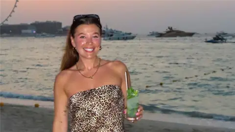 Isobel Perl Isobel on the beach with a cocktail, standing in front of the sea and a big wheel. It is dusk.