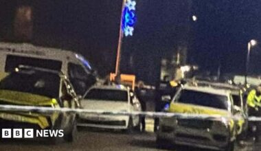 Several police cars and officers on a street in the dark, behind police tape.