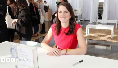 Tatiana Schlossberg with long brown hair and wearing a red short-sleeved dress, sits behind table where her book  is stood up with hands crossed holding a pen