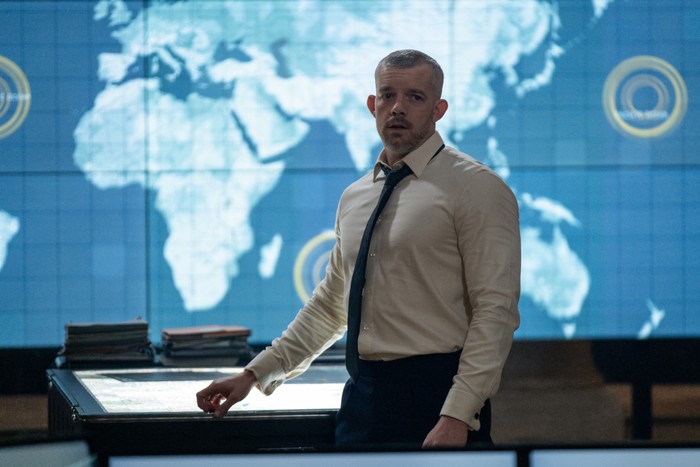 Russell Tovey as Barclay in The War Between the Land and the Sea. He's dressed in a suit anf is standing in front of a map of the world.
