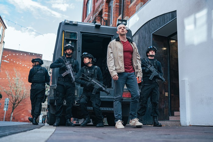 Russell Tovey as Barclay Pierre-Dupont in The War Between The Land And The Sea. He is stood in front of a black truck, and there are four officers wearing black, holding guns behind him.