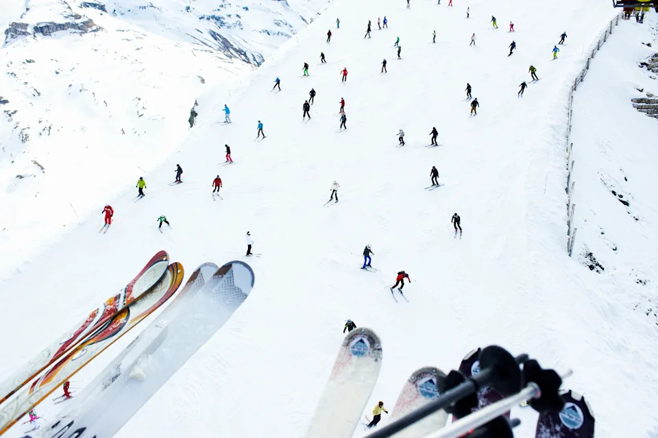 People skiing down a snowy mountain slope, with ski poles and skis visible in the foreground, capturing a busy and vibrant ski resort scene