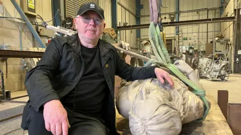 A man sits on a workshop table and places his hand on a sculpture of Lemmy, which is lying on the table next to him.