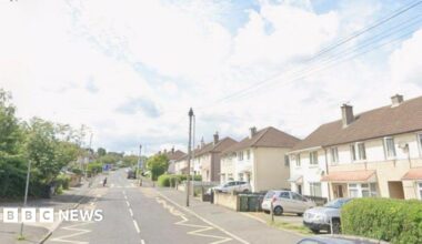 A suburban road with semi-detached houses on one side. Telegraph poles, parked cars and other street furniture is visible.
