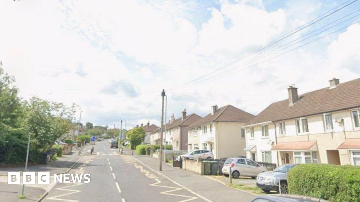 A suburban road with semi-detached houses on one side. Telegraph poles, parked cars and other street furniture is visible.