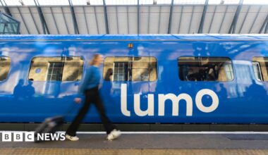A woman dressed in a blue top, dark trousers and white shoes wheeling a dark suitcase along a train platform with a blue train displaying the word Lumo in white letters on the side. The woman is blurred, suggesting she is moving.