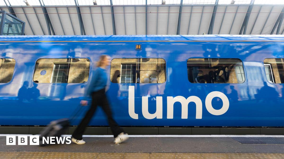 A woman dressed in a blue top, dark trousers and white shoes wheeling a dark suitcase along a train platform with a blue train displaying the word Lumo in white letters on the side. The woman is blurred, suggesting she is moving.