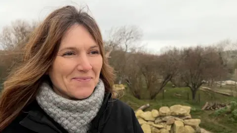 Lucie has long brown hair and is wearing a black jacket and a thick grey scarf. She is looking to the right, at the reporter who is out of frame. In the background is the bears' large enclosure, with rocks, bare trees and grass. 