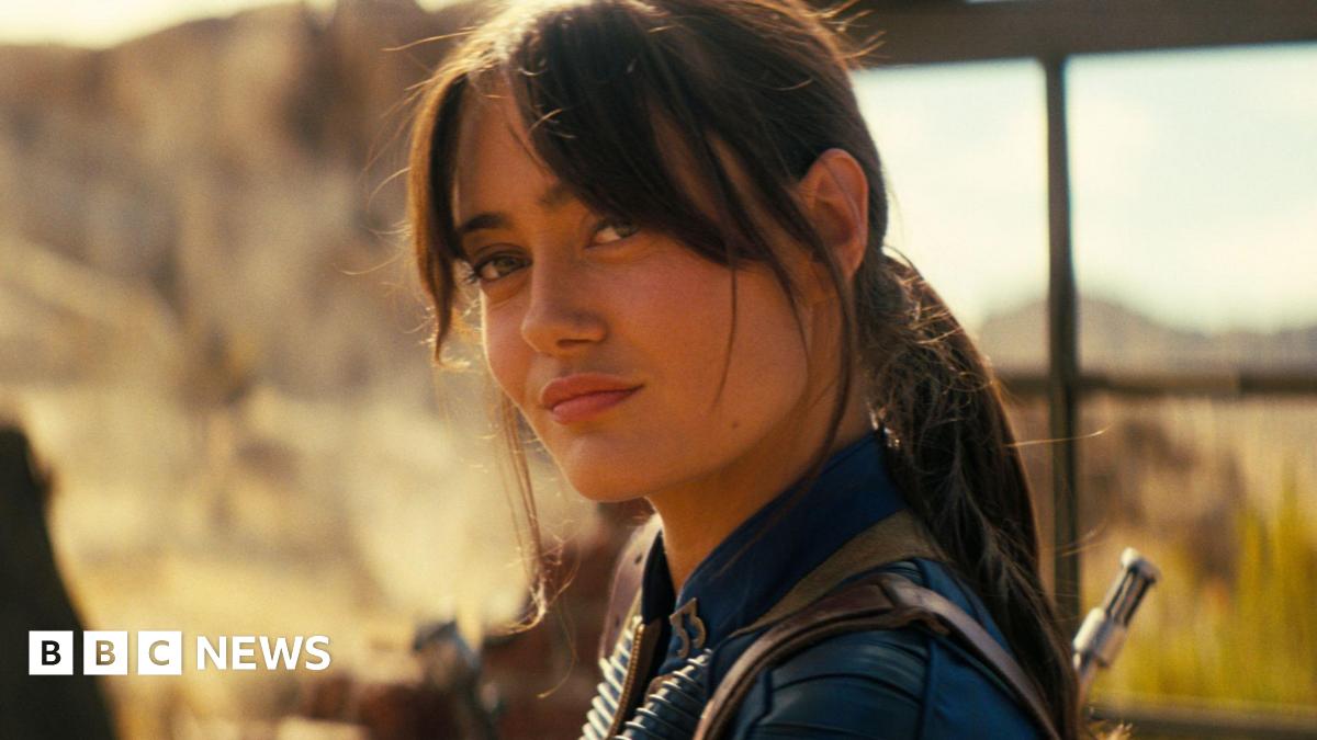 A shot from Fallout shows Ella Purnell in her blue vault suit, hair tied back, looking quizzically at something in the distance. She's surrounded by desert and bathed in a dusky yellow sunlight.