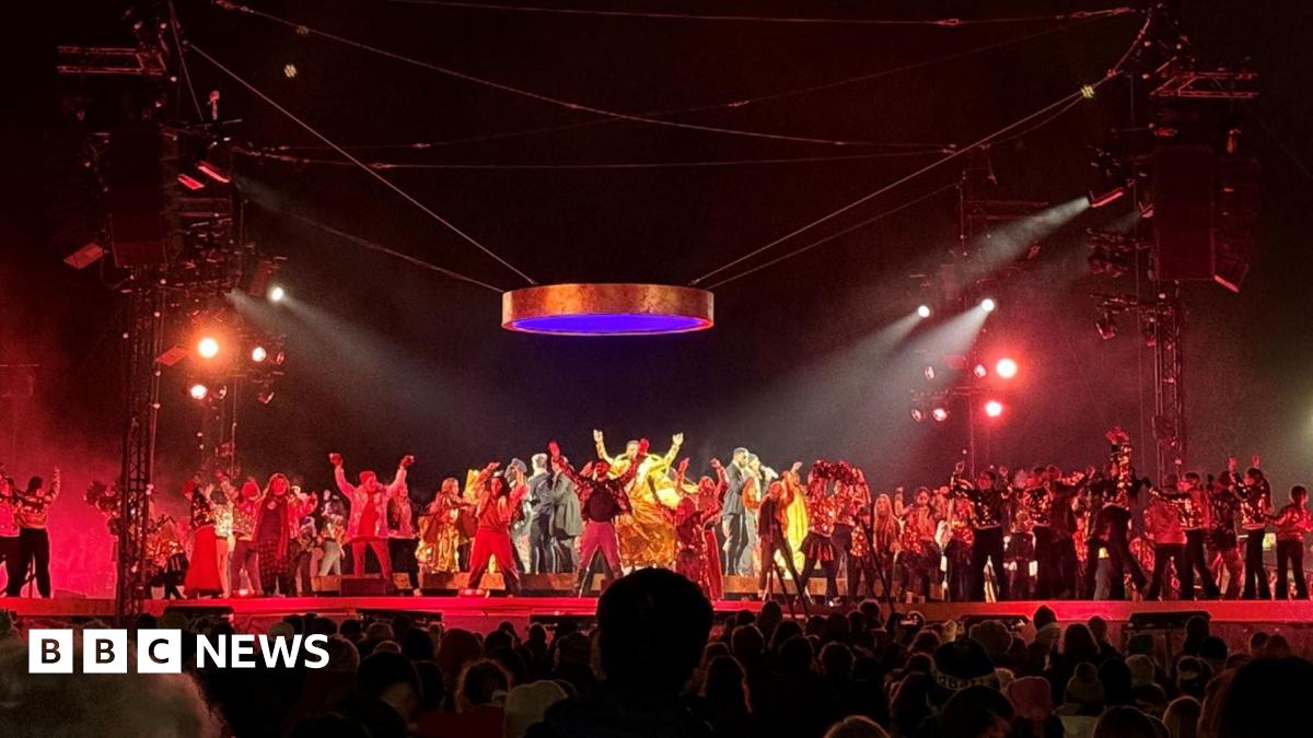 A large crowd of people spanning across a park. There are several trees in the background with hanging lights illuminating the early evening darkness.