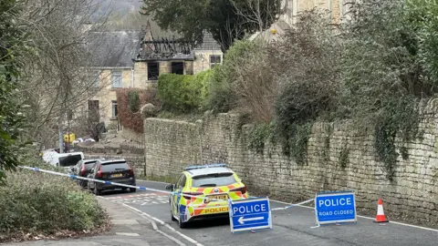 A police car blocks off a road in front of 'Police Do Not Cross tape' A row of terraced properties can be made out behind a wall. One of the properties has been badly damaged by a fire, with its roof almost entirely missing with only the bare structure visible.