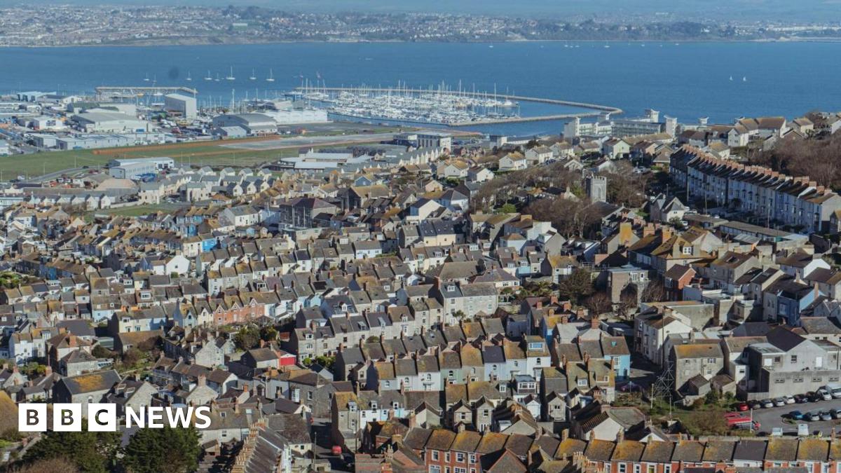 Aerial view across Portland looking towards Weymouth. In the foreground, taking up most of the photo, is the town of Fortuneswell, consisting of mostly terraced houses built on a hill. Beyond the houses is Portland Port and Portland Habour and a large marina full of small boats. On the other side of the harbour is the Wyke Regis area of Weymouth.