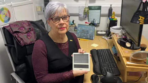 Cherry Cantrell holds a small, white light box in her hand as she sits at her desk. She is looking at the camera with a neutral expression on her face. She has short hair with a purple hue and is wearing a purple lace top and a black waistcoat. In front of her is a desktop computer.
