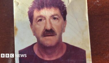 A passport-style creased photograph of a man with dark hair and a moustache. He is staring into the camera.