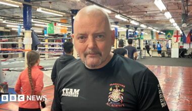 Den Doyle, a man with a grey goatee beard, stands in a boxing gym wearing a t-shirt saying Gladiators Boxing Academy. Behind him a number of people can be seen running around the room. Various colourful flags hand from the ceiling