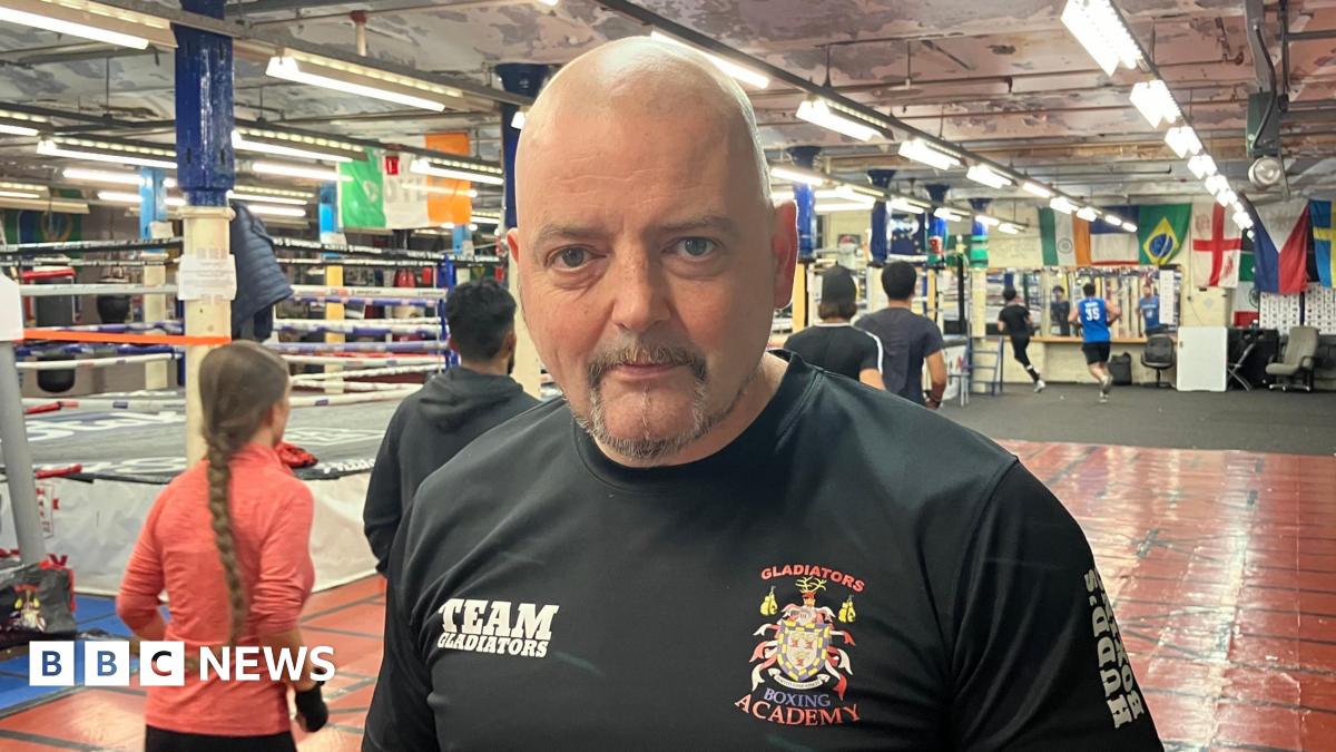 Den Doyle, a man with a grey goatee beard, stands in a boxing gym wearing a t-shirt saying Gladiators Boxing Academy. Behind him a number of people can be seen running around the room. Various colourful flags hand from the ceiling