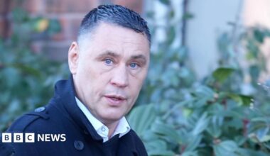 A man with short black hair wearing a black jacket. He is walking past a hedge and looking into the camera to his right. He look surprised.