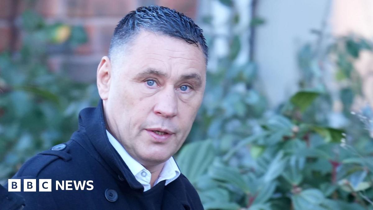 A man with short black hair wearing a black jacket. He is walking past a hedge and looking into the camera to his right. He look surprised.