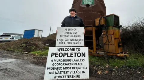 An elderly man stands on muddy and rocky land holding two white signs. The first sign reads: "As seen on Jeremy Vine Tuesday 16 March A brilliant interviewer but a lousy dancer." The second sign, which is slightly larger and resting on his feet, reads: "Welcome to Peopleton a murderous lawless godforsaken place probably, most definitely the nastiest village in Worcesteshire."