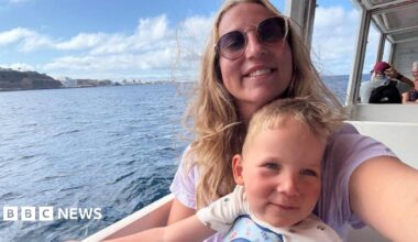 Edward stands in a blue, black and grey wetsuit next to a swimming pool with two people in it. He has dark blonde wavy hair.