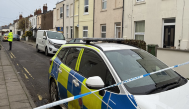A police cordon on a residential street with a police car behind blue and white evidence tape and an officer standing guard halfway down the road