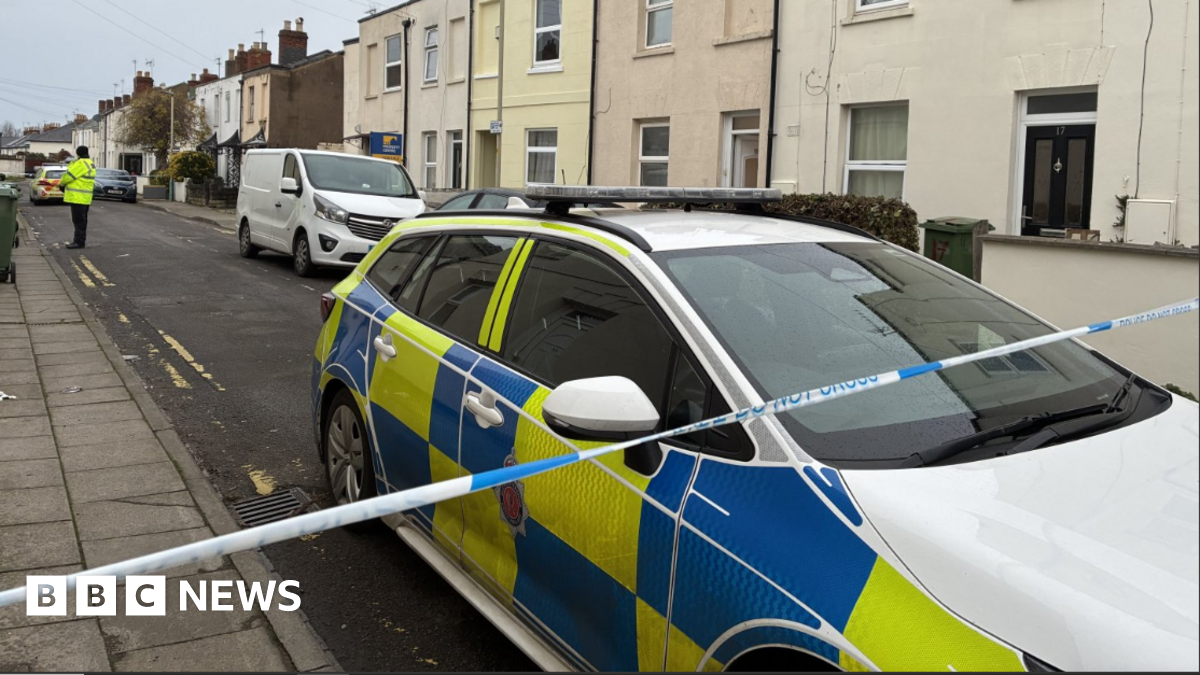 A police cordon on a residential street with a police car behind blue and white evidence tape and an officer standing guard halfway down the road