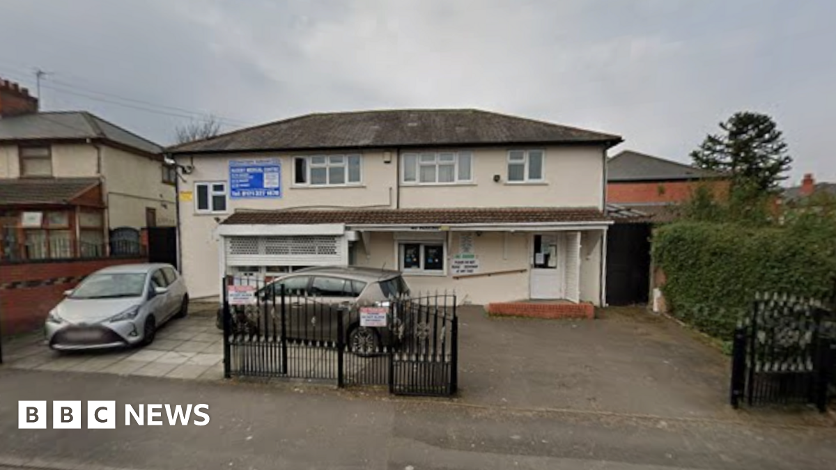 General view of the entrance of Naseby Medical Centre in Birmingham. A cream coloured building with a small driveway outside. Two cars are parked in the driveway which has some iron gates across part of it.