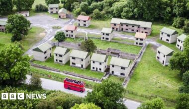 An aerial picture of what the village looks like today, with a small cluster of empty brick houses with tin roofs and no windows or doors. It is a stark and bare street and all the houses look identical. At the bottom there is a red double-decker tour bus driving along the road.