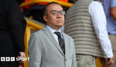 Jeff Shi at Molineux in a grey suit, a shirt and tie and dark-rimmed glasses, looking in the direction of the camera