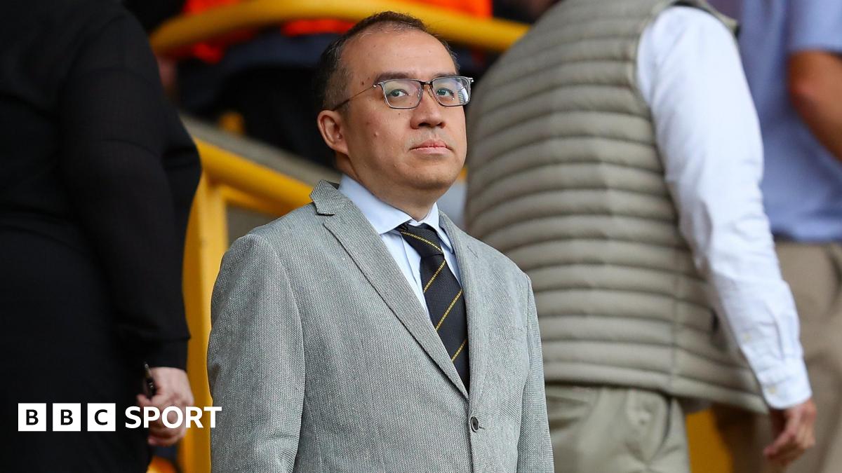 Jeff Shi at Molineux in a grey suit, a shirt and tie and dark-rimmed glasses, looking in the direction of the camera