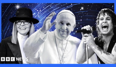 D’Angelo is performing on stage, wearing a wide-brimmed black hat and a black leather vest over a deep V-neck top. A silver chain necklace is visible around the neck, and the left arm shows a detailed tattoo design. He is positioned near a microphone with a black foam cover. The background features a warm orange gradient, creating a dramatic and vibrant concert atmosphere.