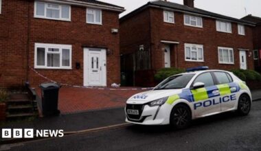 A police car is parked on the road outside a house, where police crime tape can be seen cordoning off the brick-paved driveway. The house has a white door and a white-framed front lower window.