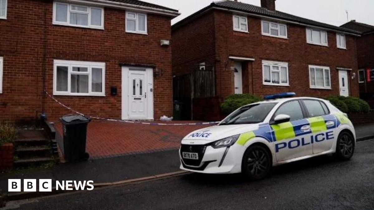 A police car is parked on the road outside a house, where police crime tape can be seen cordoning off the brick-paved driveway. The house has a white door and a white-framed front lower window.