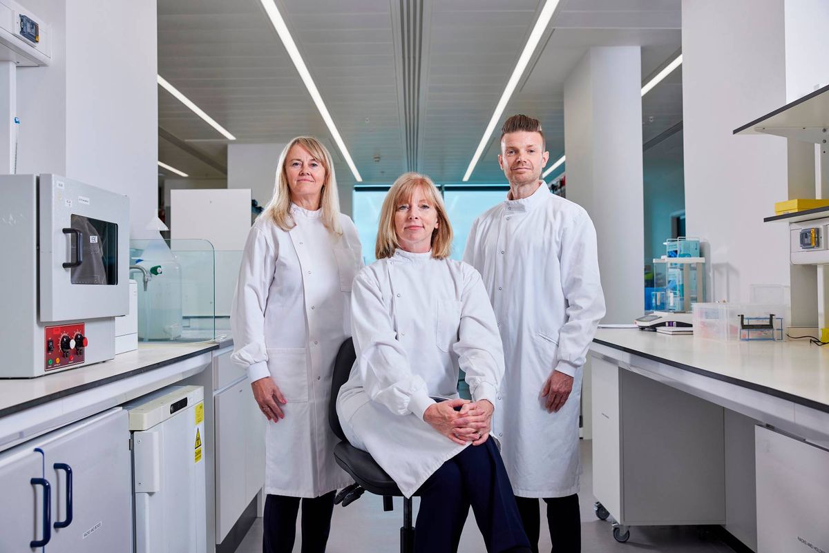 Three individuals in white lab coats are standing in a modern laboratory, possibly engaged in scientific research or discussion. The setting features contemporary laboratory equipment and white countertops.
