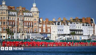 Line of runners in red Santa outfits making their way along Weymouth Beach with the sea to the right and buildings along the left.