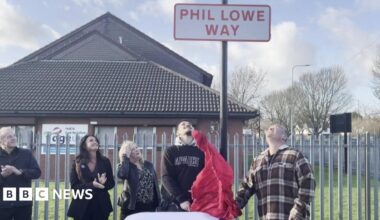 Five people stand underneath a road sign. It is in red letters against a white background and says Phil Lowe Way. Underneath are members of his family, including his son, who has just pulled down a red sheet that was covering the sign.