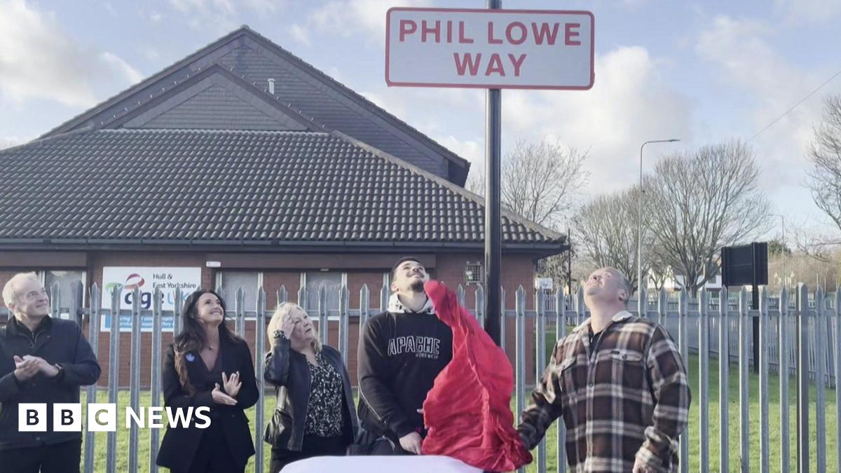 Five people stand underneath a road sign. It is in red letters against a white background and says Phil Lowe Way. Underneath are members of his family, including his son, who has just pulled down a red sheet that was covering the sign.