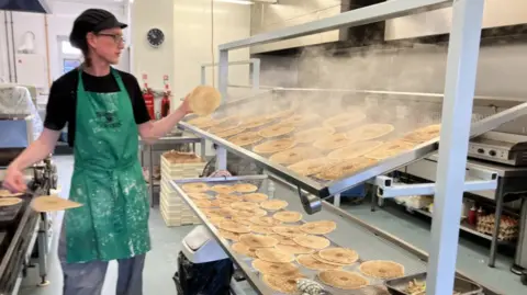 Large racks of circular oatcakes, with steam rising from them. A worker is adding more to the racks 
