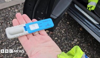 A police officer's hand holding a blue and grey drug testing kit.