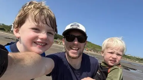 Andrew Andrew takes a selfie on the beach on a gloriously sunny day with blue skies. His sons Joey and Tommy stand in front of him in their mini wetsuits.