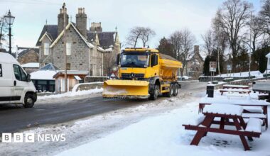 A yellow gritting lorry fitted with a snow plough travels down a snowy street in the picturesque village of Braemar.