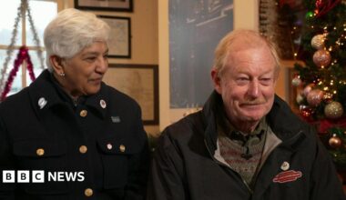 Chandra and Richard Law sit inside a railway station next to a Christmas tree decorated with pink and gold baubles. Chandra has short grey hair and is wearing a wool coat with brass buttons. She is smiling at Richard who is wearing a blue coat and a green jumper. He has light-coloured hair and a moustache.