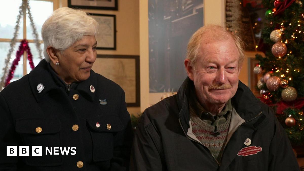 Chandra and Richard Law sit inside a railway station next to a Christmas tree decorated with pink and gold baubles. Chandra has short grey hair and is wearing a wool coat with brass buttons. She is smiling at Richard who is wearing a blue coat and a green jumper. He has light-coloured hair and a moustache.
