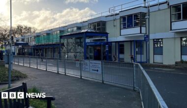 Exterior of QEH hospital front. Modern building painted in blue and yellow. Railings are opposite with a road dissecting the two. Some scaffolding is on the building.