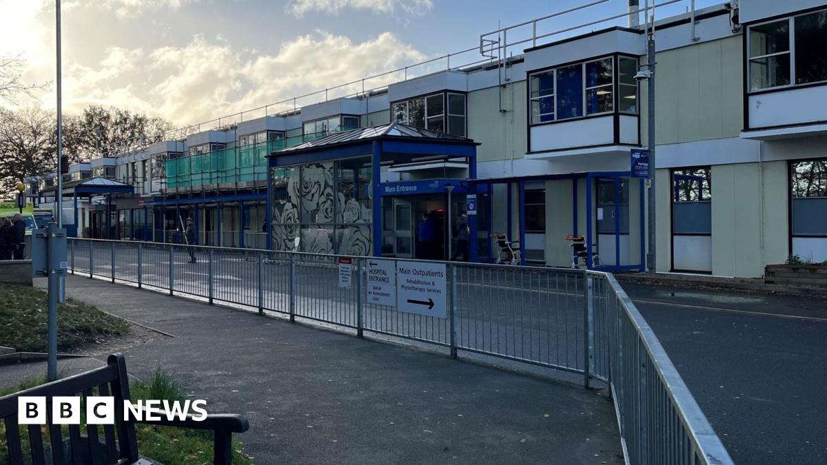 Exterior of QEH hospital front. Modern building painted in blue and yellow. Railings are opposite with a road dissecting the two. Some scaffolding is on the building.