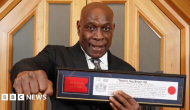 Frank Bruno holding his framed certificate showing his freedom of the City of London. His right hand is holding his fist in a boxing punch toward camera.