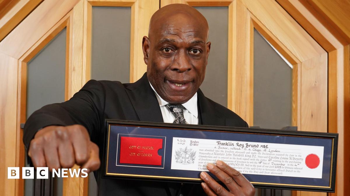 Frank Bruno holding his framed certificate showing his freedom of the City of London. His right hand is holding his fist in a boxing punch toward camera.