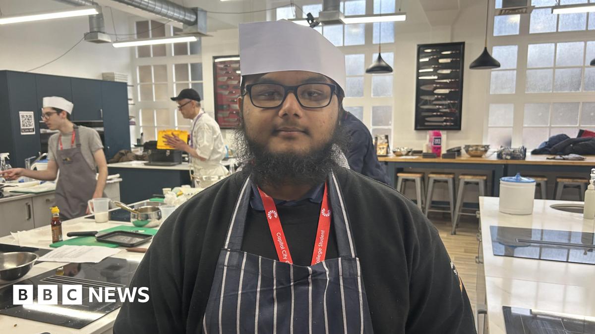 A young man in a chef's uniform looks into the camera in a busy training kitchen. He wears a white chef hat, a red lanyard, and a blue and white striped apron.