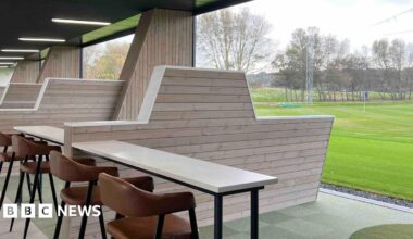 A golfing green is outside with a table and chars inside a driving range , with a large barrier between ranges under cover.