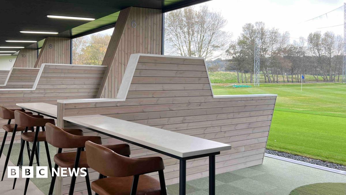 A golfing green is outside with a table and chars inside a driving range , with a large barrier between ranges under cover.